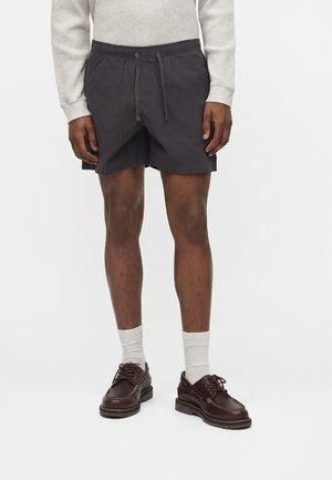 Άντρας που φοράει σκούρα σορτς με κορδόνι, κρεμ μακρυμάνικη μπλούζα με υφή βάφλας, λευκές κάλτσες και σκουρόκαφέ δερμάτινα παπούτσια με κορδόνια σε λευκό φόντο.