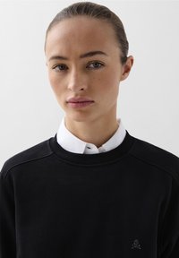 Sudadera negra con cuello redondo, puños de canalé y un pequeño logo de calavera. Camisa blanca con cuello por debajo, textura suave, diseño minimalista.