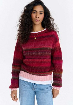 Czerwony i różowy sweter w paski z dzianiny, z prążkowanym dołem i mankietami, o miękkiej teksturze i luźnym kroju, noszony na białej koszuli.