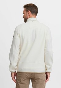 Pull en polaire blanche avec col montant, manches longues et ourlet côtelé. Présente un petit logo sur la manche gauche. Tissu texturé.
