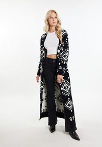 Lång svart och vit cardigan med geometriska mönster, stickad struktur, öppen front, bärs över en vit kort topp och svarta byxor.