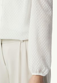Weiße strukturierte Bluse mit kariertem Muster, gerafften Bündchen und leichtem Stoff, kombiniert mit cremefarbenen, schmal zulaufenden Hosen.