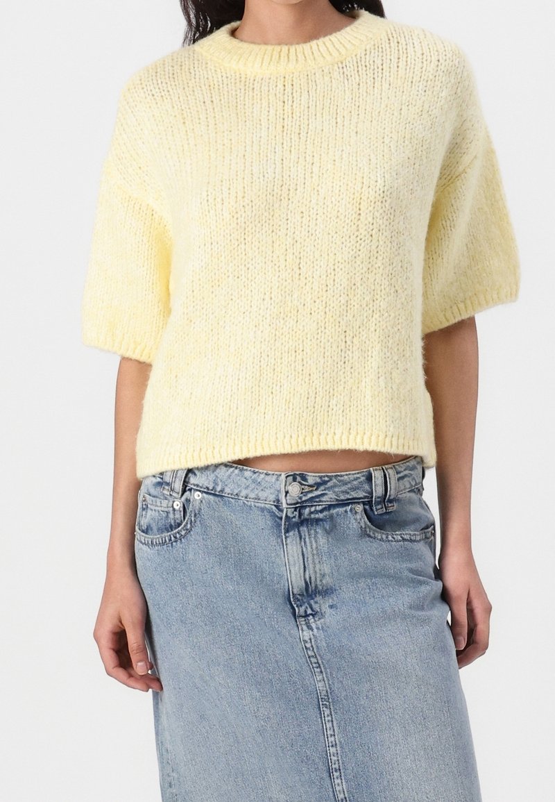 Femme portant un pull en maille jaune pâle à manches courtes et une jupe en jean bleu clair taille haute sur fond blanc.