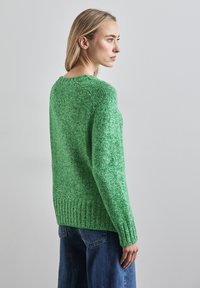 Grüner Strickpullover mit strukturierter Oberfläche und geripptem Saum, mit rundem Halsausschnitt und überschnittenen Schultern, kombiniert mit lockeren blauen Jeans.