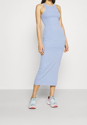 Kobieta w bezrękawowej, jasnoniebieskiej, dopasowanej sukience midi, w parze z jasnoniebiesko-różowymi sportowymi butami, stojąca na jednolitym tle.
