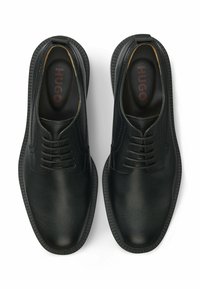 Paar schwarze Lederschnürschuhe mit strukturierter Oberfläche und dicken Gummisohlen, innen mit dem Markennamen "HUGO" sichtbar.