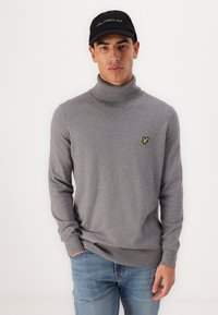 Grijze turtleneck trui van een zachte stof, met een klein geel geborduurd logo aan de linkerkant. Gecombineerd met blauwe spijkerbroeken.