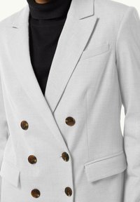 Blazer gris claro de doble botonadura con botones marrones, solapas de pico y bolsillos laterales. La tela parece texturizada, complementando un jersey negro de cuello alto.