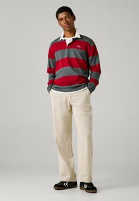 Sarkans un pelēks svītrains garo piedurkņu polo krekls ar baltu apkaklīti, apvienots ar krēma krāsas samta biksēm un melniem sporta apaviem ar baltām detaļām.