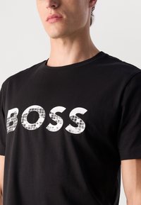 Schwarzes Baumwoll-T-Shirt mit einem großen weißen "BOSS"-Logo, das ein reflektierendes, strukturiertes Muster aufweist. Rundhalsausschnitt und kurze Ärmel.