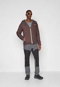 Brun isoleret jakke med mintfarvet lynlås, grå sweatshirt under, og bukser i sort og grå med farveblokdesign samt mørke sko.