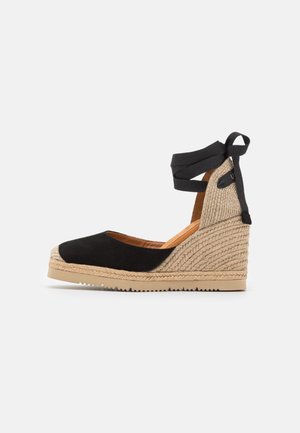 Espadrille compensée en suède noir avec détails en jute, talon ouvert et strap au niveau de la cheville. Texture lisse et design à bout rond, hauteur de talon de 7,5 cm.