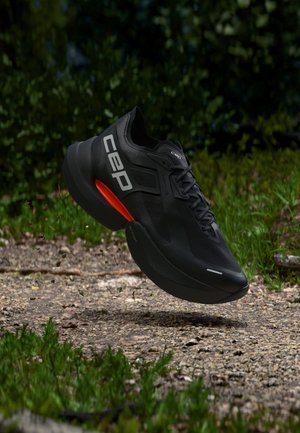 Black running shoe with white "CEP" logo and red detail mid-air over gravel path surrounded by green foliage.