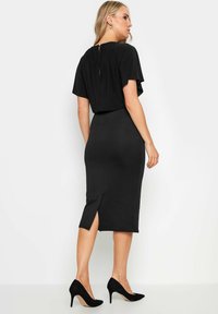 Robe midi noire avec une jupe crayon ajustée, des manches courtes à ailettes lâches et une ouverture en forme de clé au dos. Comprend une fente sur le côté.