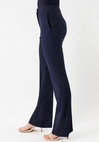 Donna che indossa pantaloni svasati su misura color blu navy e sandali beige con tacco, in piedi contro uno sfondo bianco uniforme.