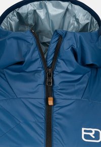 Blaue isolierte Jacke mit hohem Kragen, Reißverschluss und gestepptem Muster. Mit kontrastierender silberner Innenseite und Logoakzent.