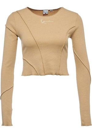 Top court beige à manches longues avec un design de couture asymétrique et une signature blanche brodée près de l'encolure.