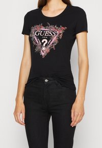 Černé bavlněné tričko s krátkým rukávem, které zdobí grafický design květinového trojúhelníku se textem "GUESS U.S.A." a centrálním otazníkem.