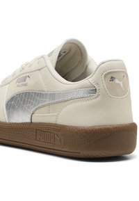 Zapatillas Puma Palermo en cuero crema con un logo lateral plateado texturizado. Suela de goma marrón con un patrón texturizado. Diseño minimalista.