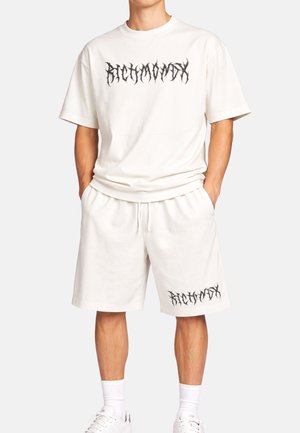 Uomo che indossa un completo composto da T-shirt oversize bianca e pantaloncini con scritta stilizzata nera sul petto e sulla gamba sinistra, abbinato a sneakers bianche e calzini.