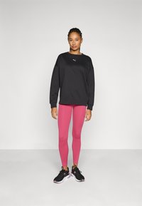 Sudadera negra con puños acanalados, leggings rosas y zapatillas deportivas negras. La sudadera cuenta con un logo blanco; los leggings tienen una textura suave.