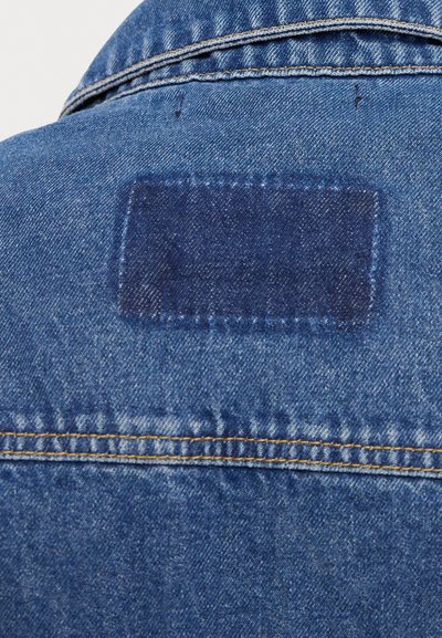 Denimjakke med et rektangulært marineblått stoffmerke bak. Har klassisk blå denimtekstur og synlig gyllen søm på sømmene.