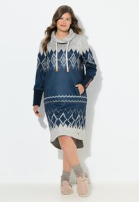 Vrouw in een blauw en grijs gebreid jurk met een col, lichte bruine sokken en beige instappers, staand tegen een witte achtergrond.