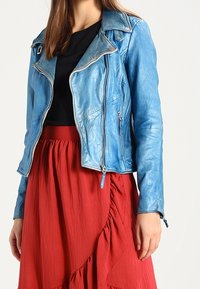 Frau trägt eine blaue Lederjacke, ein schwarzes Oberteil und einen roten, rüschierten Rock, der an Hals und Oberschenkel endet, vor einem schlichten weißen Hintergrund.