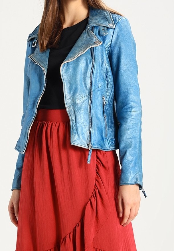 Frau trägt eine blaue Lederjacke, ein schwarzes Oberteil und einen roten, rüschierten Rock, der an Hals und Oberschenkel endet, vor einem schlichten weißen Hintergrund.