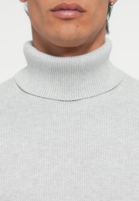 Svijetlosiva rebrasta turtleneck džemper sa prikladnim dizajnom i teksturiranom površinom. Ima preklopni ovratnik i detalje sa besprijekornim uzorkom.