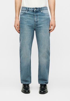 Férfi világoskék egyenes szárú farmernadrágban, fekete elegáns cipővel és beletűrt fehér inggel, fehér háttér előtt áll.