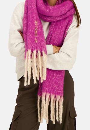 Personne portant une écharpe à franges rose vif avec le texte "MY JEWELLERY", un pull gris clair, un pantalon cargo foncé et un bracelet en or.