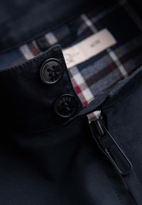 Gros plan sur le col d'une veste noire avec deux boutons noirs et une fermeture éclair, montrant une doublure à carreaux bleue et blanche à l'intérieur.