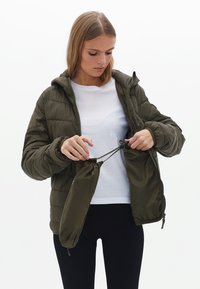 Olijfgroene pufferjack met capuchon, voorzien van een gewatteerd ontwerp en elastische manchetten. Model past de trekkoord van de binnenzak aan.