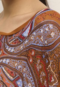 Vzorovaný top s hnědým podkladem, který má složité modré a červené paisley vzory. Široký výstřih a jemná textura.