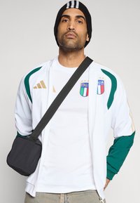 Valkoinen vetoketjullinen takki, jossa vihreät yksityiskohdat ja kultaiset raidat, valkoinen t-paita, jossa italialainen vaakuna, musta pipo ja pieni musta olkalaukku.