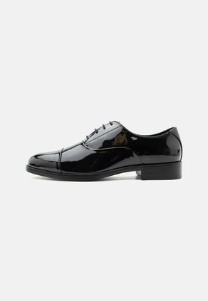 Zwarte lakleren herenschoenen met gesloten vetersluiting en een lage hak, zijaanzicht op een witte achtergrond.
