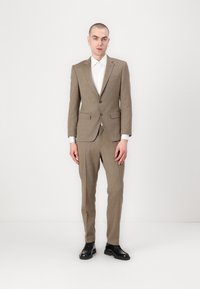 Completo in tessuto di lana a motivo check marrone, che presenta un blazer aderente con due bottoni e pantaloni coordinati, abbinati a una camicia bianca e scarpe nere.