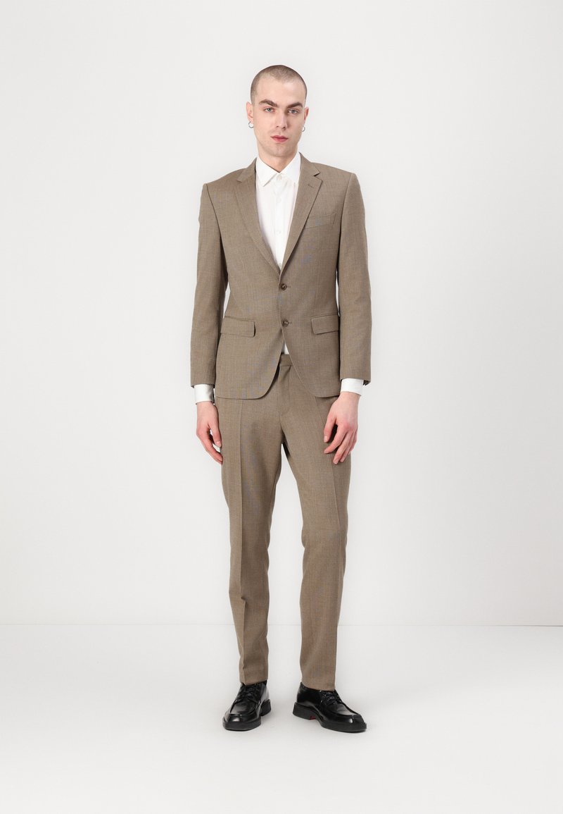 Completo in tessuto di lana a motivo check marrone, che presenta un blazer aderente con due bottoni e pantaloni coordinati, abbinati a una camicia bianca e scarpe nere.