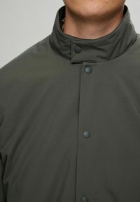 Mørkegrønn, vannavstøtende jakke med høy krage, trykknapplukking og glatt tekstur. Ingen mønstre eller ekstra detaljer synlige.