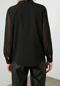 Blusa nera con maniche lunghe trasparenti, polsini con bottone e orlo arrotondato, indossata con pantaloni in pelle nera a vita alta.