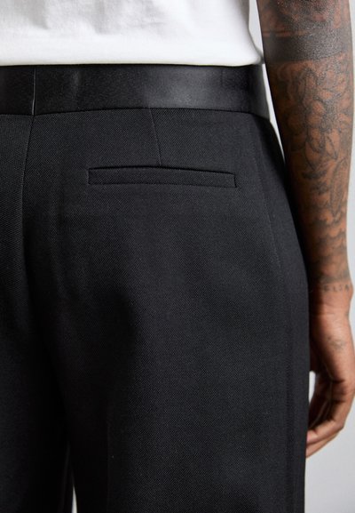 Pantalons noirs sur mesure, en tissu texturé, taille ajustée avec une ceinture noire et détail de poche arrière sur le côté gauche.