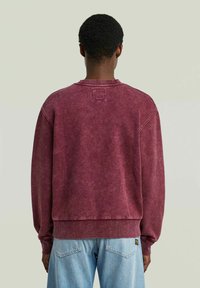 Felpa bordeaux con collo a girocollo a costine, spalle basse e un'etichetta quadrata sul retro, abbinata a jeans azzurri.