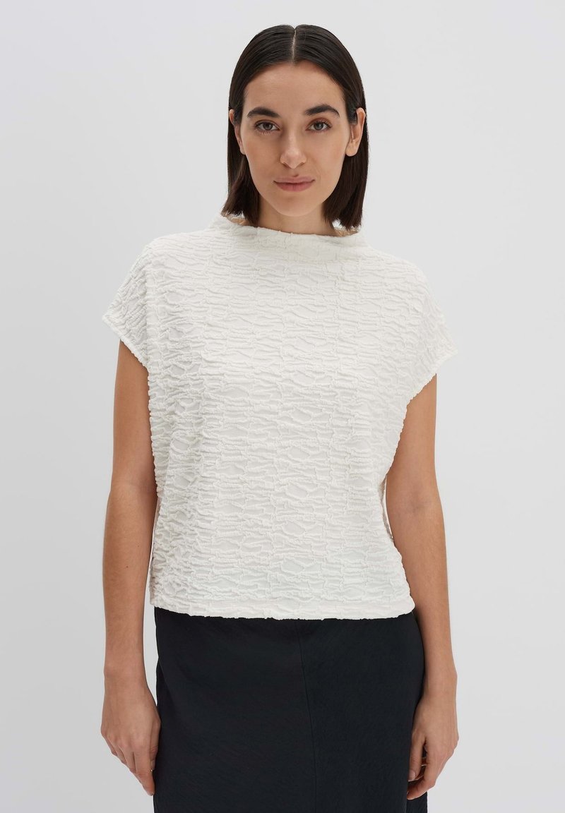 Top bianco testurizzato con collo alto e maniche corte, caratterizzato da un motivo ondulato e una vestibilità comoda, abbinato a una gonna scura.