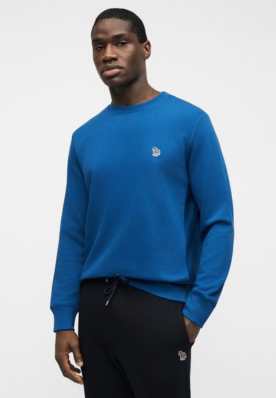 Άνδρας που φοράει μπλε φούτερ με μακριά μανίκια και μαύρο παντελόνι, και στα δύο υπάρχει ένα μικρό πολύχρωμο λογότυπο ζώου στο στήθος και στο μηρό.