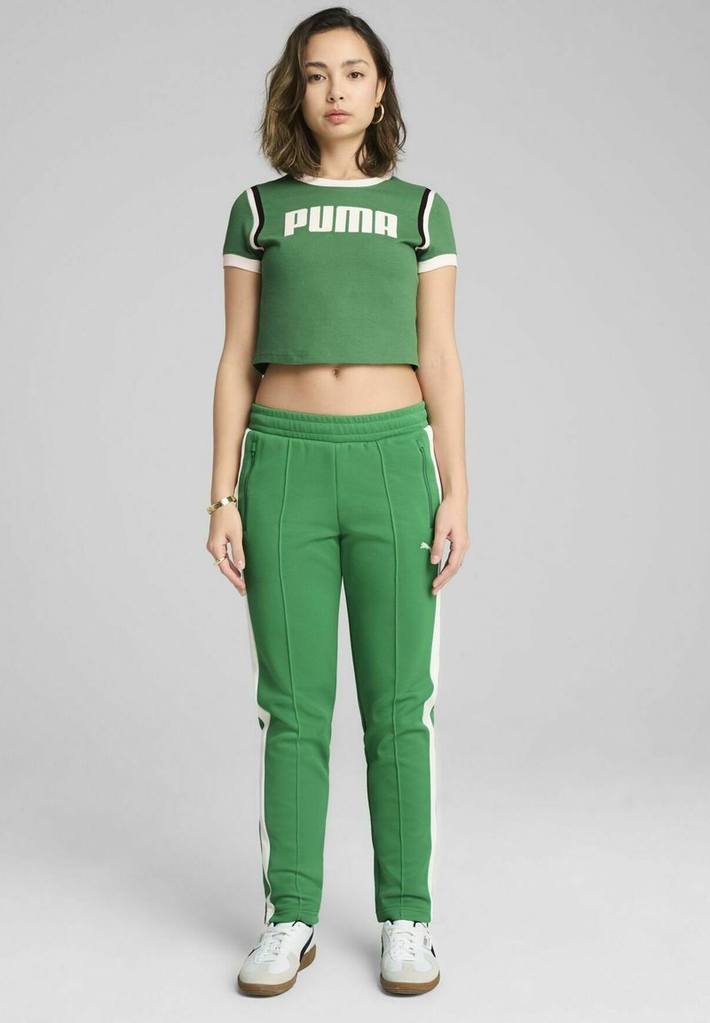 Zeleni skrajšani majica s kratkimi rokavi z logotipom "PUMA", v kombinaciji z zelenimi trenirkami, ki imajo bele stranske črte. Model nosi bele superge.