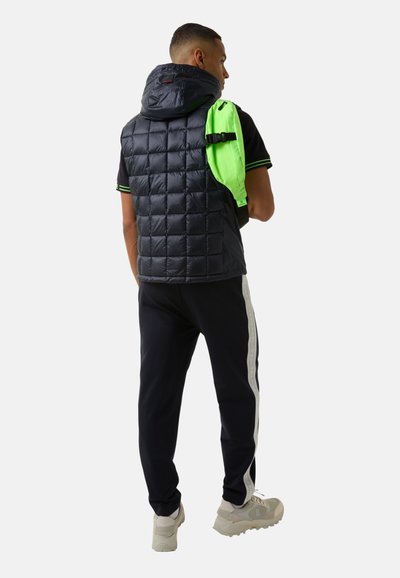 Schwarze gesteppte Steppweste mit Kapuze, kombiniert mit einem schwarzen Shirt und navyblauen Hosen mit weißen Seitenstreifen, sowie einem leuchtend grünen Rucksack.