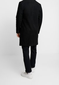 Cappotto lungo nero con una texture liscia, caratterizzato da un design con cucitura centrale sul retro. Abbinato a pantaloni scuri e sneakers bianche.