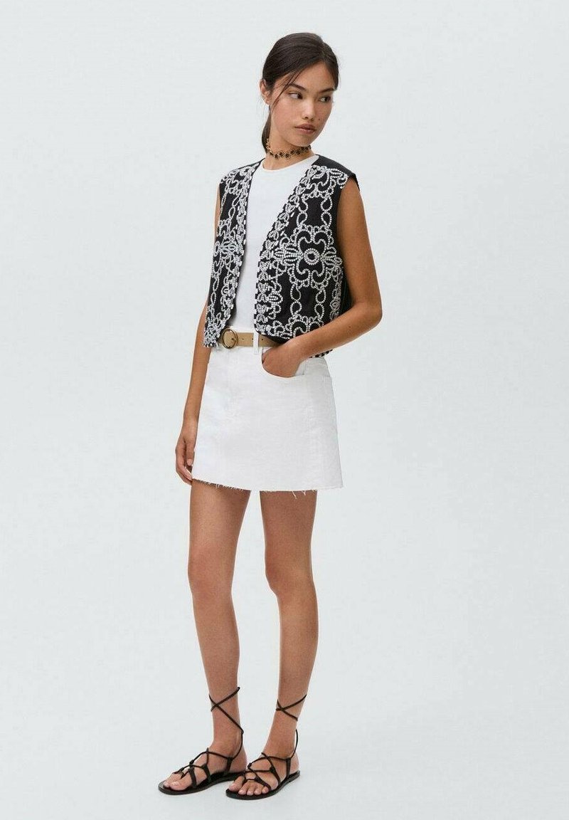 Un gilet noir et blanc brodé de motifs floraux sur un débardeur blanc, associé à une jupe en denim blanche et des sandales noires à lanières.