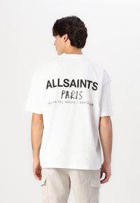 White cotton t-shirt with black text on the back reading "ALLSAINTS PARIS" and "ACROSS ALL AREAS." Relaxed fit, short sleeves.